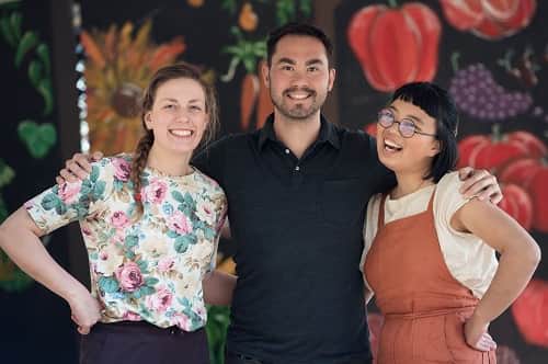 Joyce Liao, Meryn Corkery and Colin Dring stand smiling and arm-in-arm in front of a painted mural of vegetables.