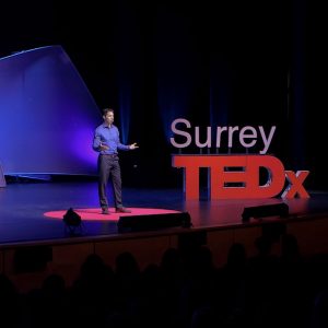 Dr. Kai Chan presenting on stage with a purple backlight and a large red "Surrey TEDx" sign on the ground.