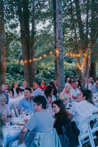 Diners sitting at long tables outdoors between trees under twinkly lights.